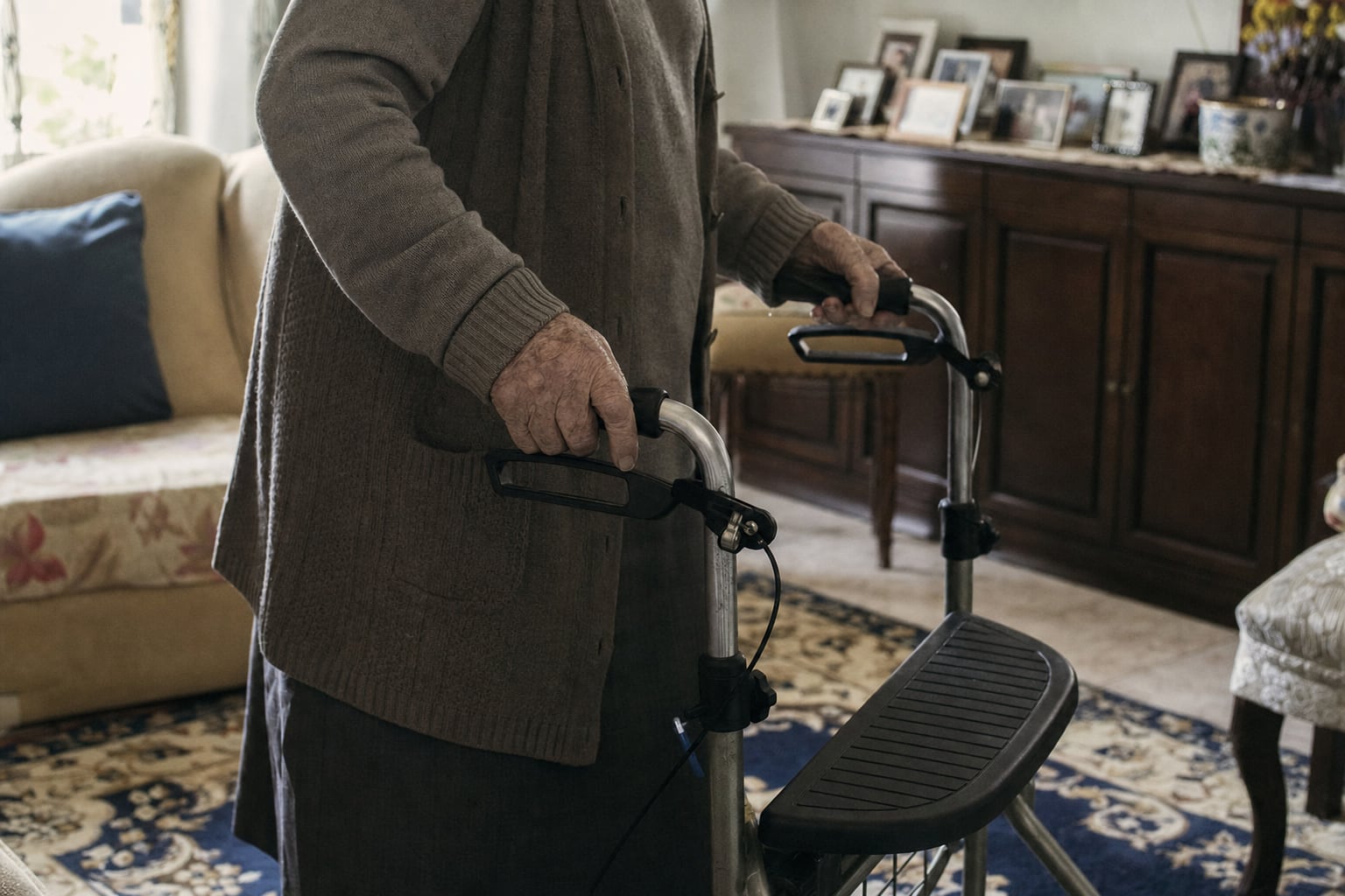 Hände einer älteren Frau auf den Griffen eines Rollators in einem hellen Wohnzimmer